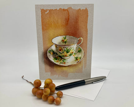 Teacup and saucer with floral design on a textured card, with a pen and fruit on a white surface.