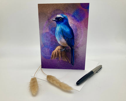 Greeting card with a blue bird illustration on a purple background, accompanied by a pen and decorative plant on a white surface.