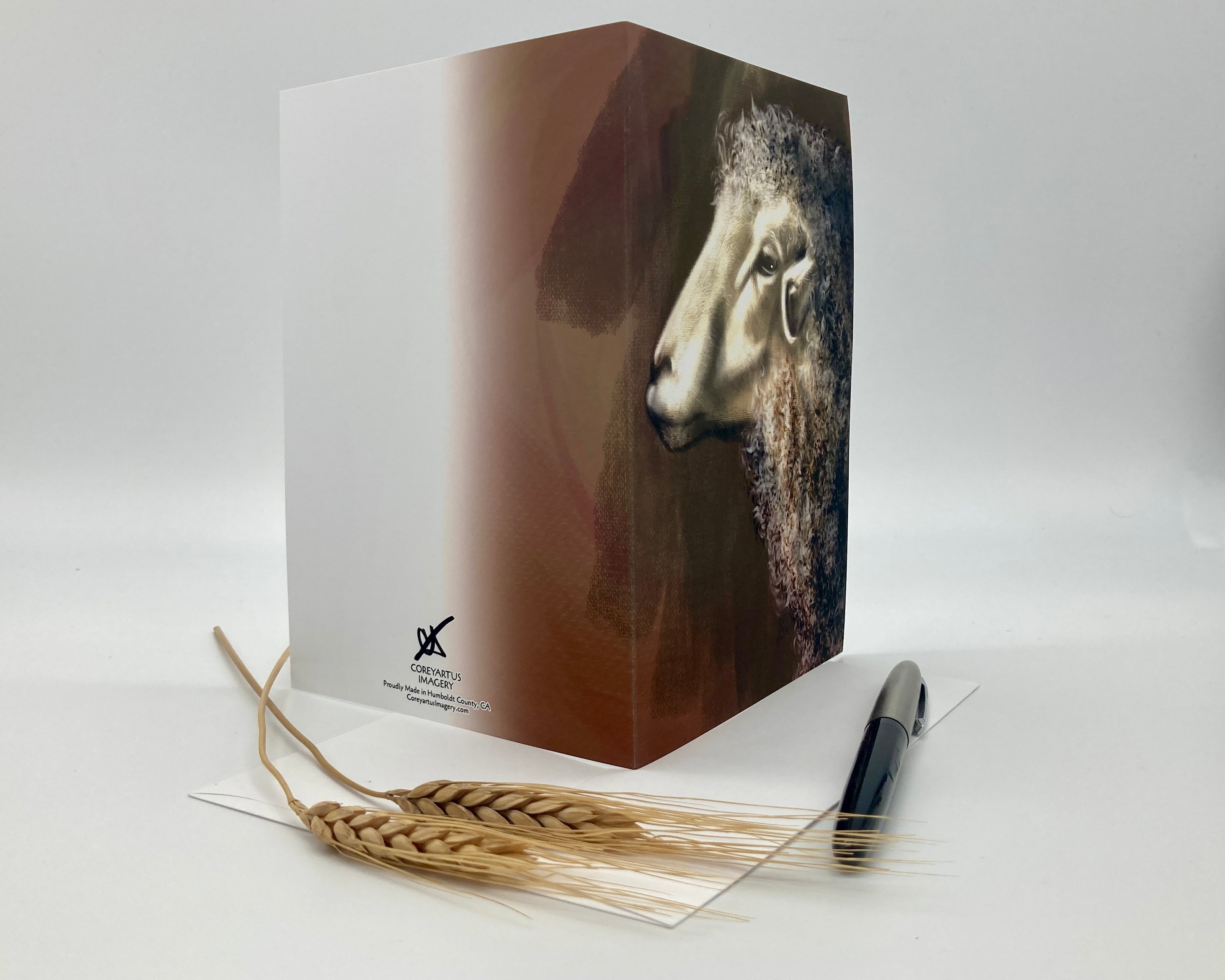Open greeting card with a sheep design on the cover, accompanied by wheat stalks and a pen on a white background.