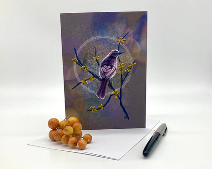 Greeting card with a bird illustration on a branch with flowers, placed on a white surface with a pen and small round objects.