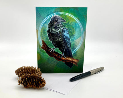Greeting card with a raven design on a white surface with a pen and pine cone.