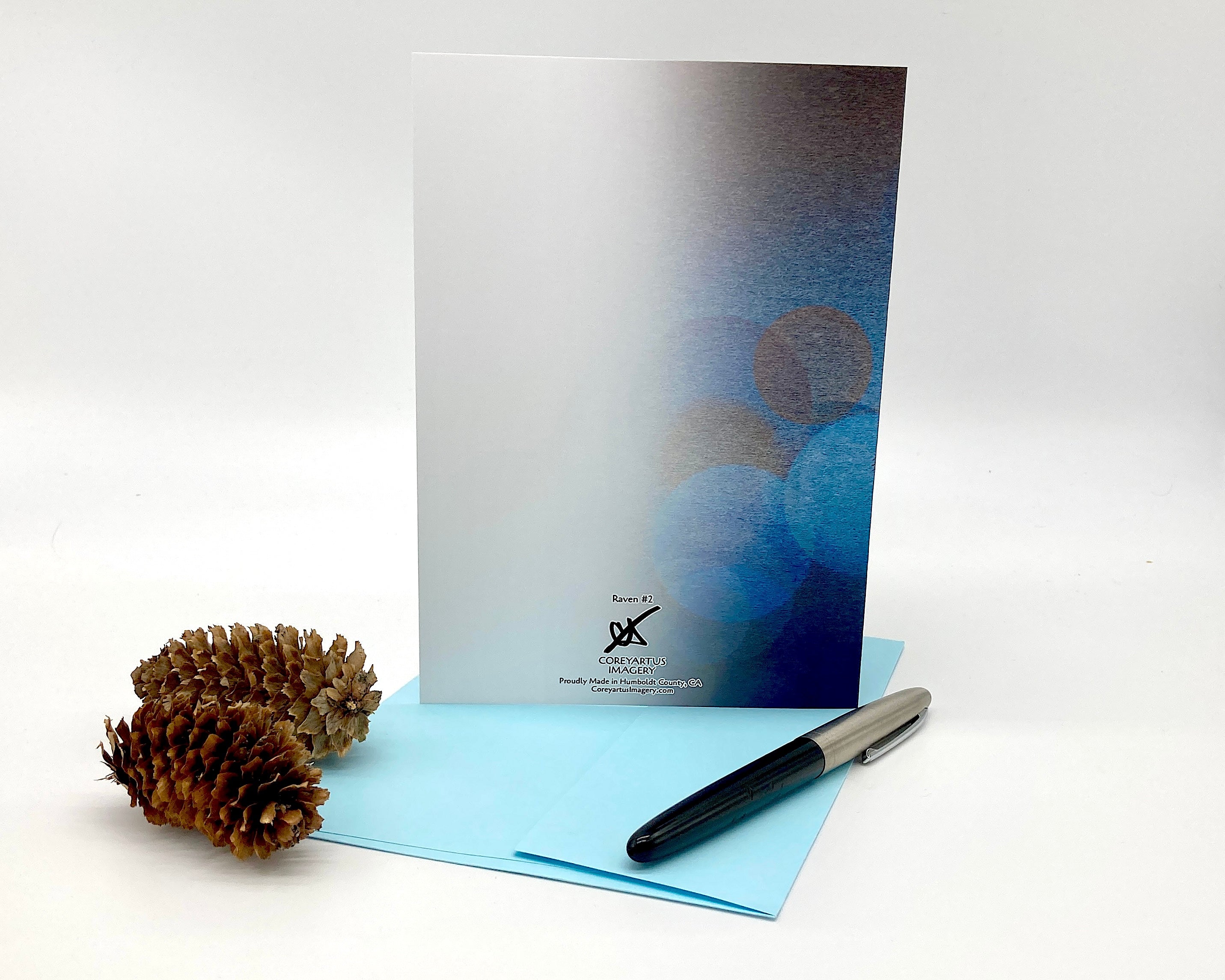 Greeting card with blue watercolor design, pen, and pine cone on a white background