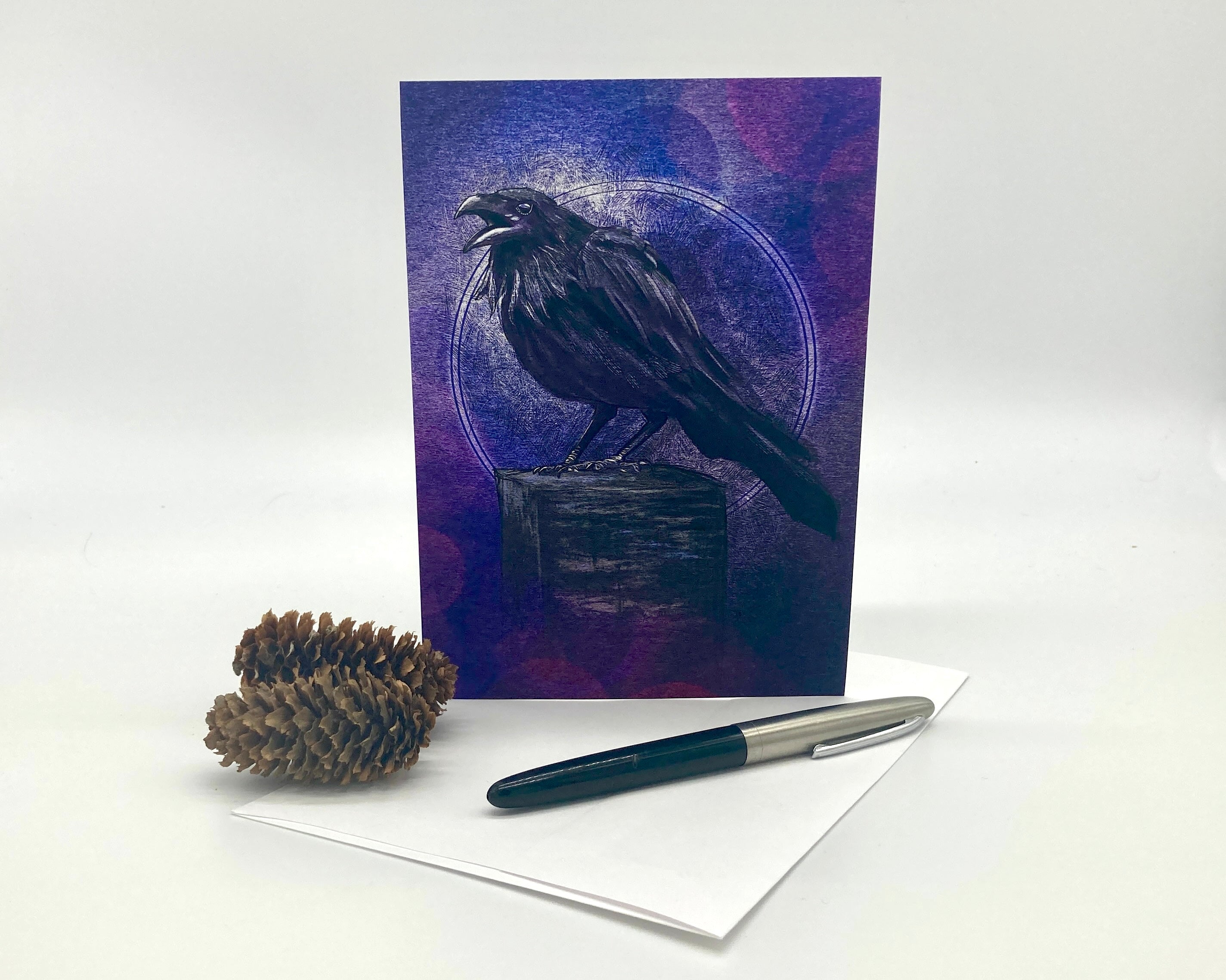 Greeting card with a raven design on a purple background, accompanied by a pen and pine cone on a white surface.