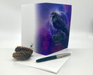 Greeting card with a raven design on a white surface with a pen and pine cone.