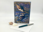 Decorative greeting card with a bird design on a white surface with a pen and shell.