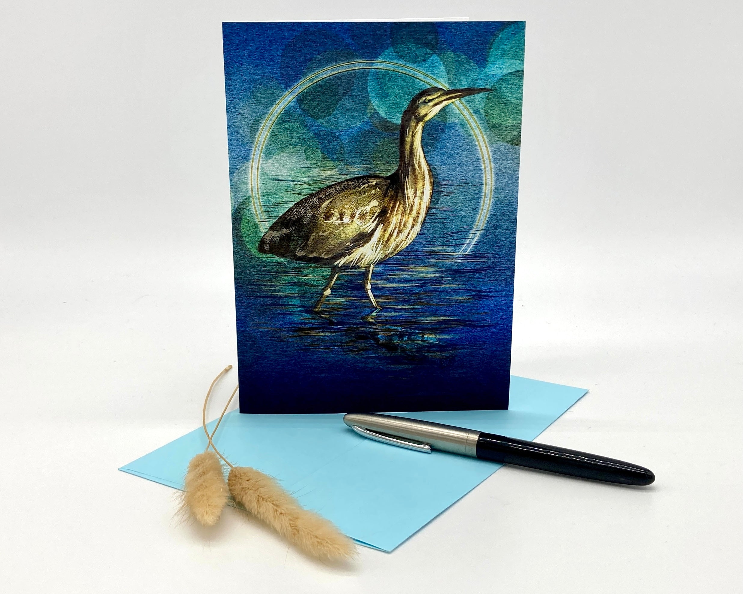 Greeting card with a bittern design on a blue background, accompanied by a pen and decorative item.