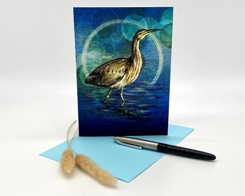 Greeting card with a bittern design on a blue background, accompanied by a pen and decorative item.