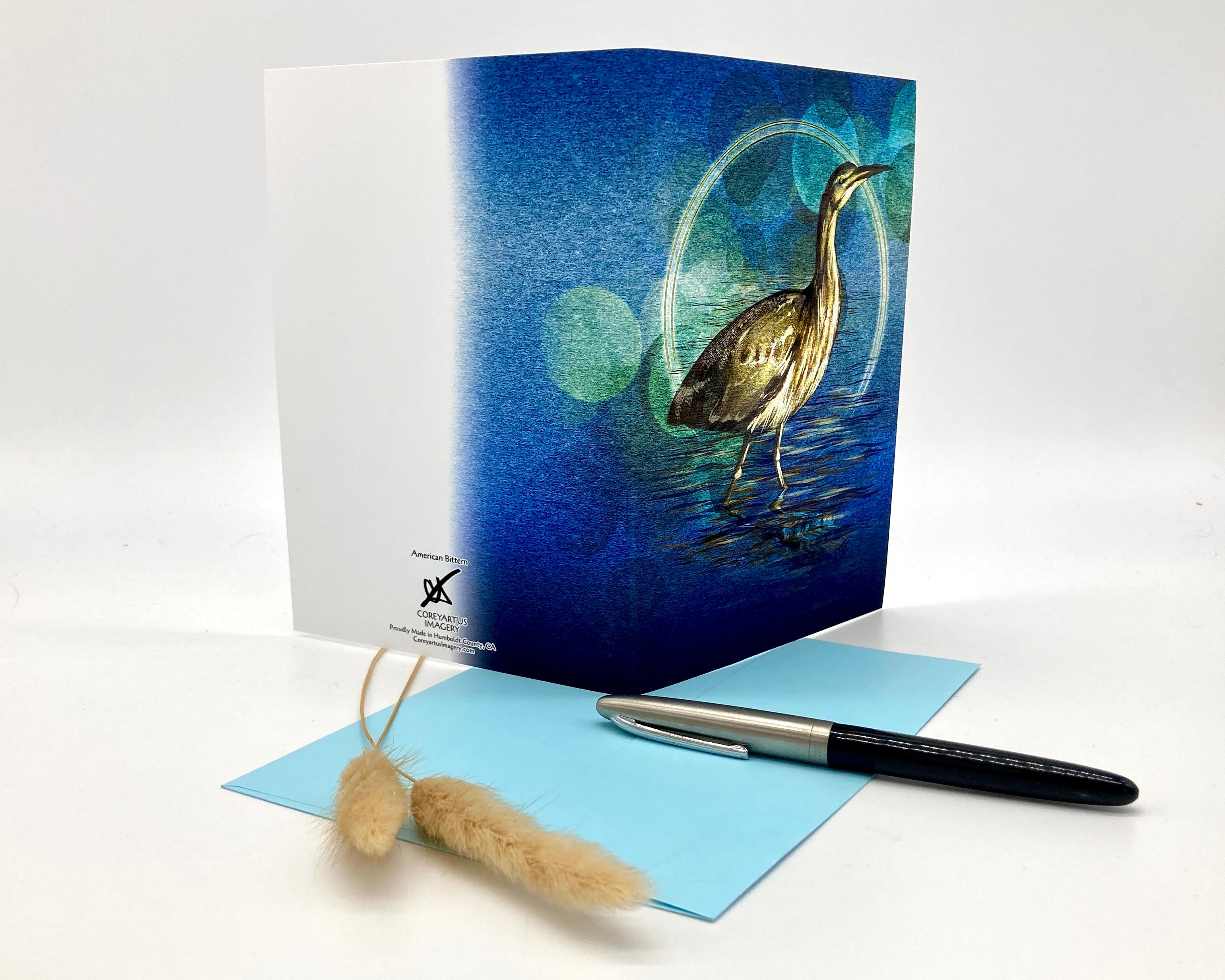 Greeting card with a heron design on a blue background, accompanied by a pen and small object.