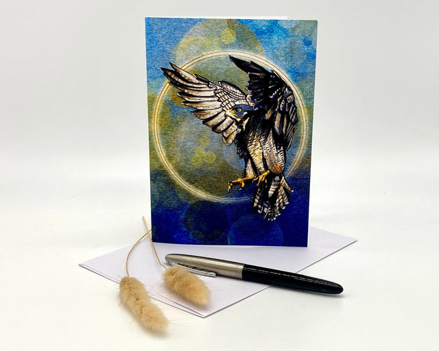 Greeting card with an eagle design on a blue background, accompanied by a pen and feather on a white surface.