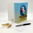 Greeting card with a colorful bird illustration on a white surface with a pen and dried plants.