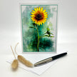 A greeting card featuring a watercolor-style yellow sunflower on a textured green and teal background, displayed upright with an envelope and a black-and-silver pen on a white surface. Two dried bunny tail grass stems are placed to the left.