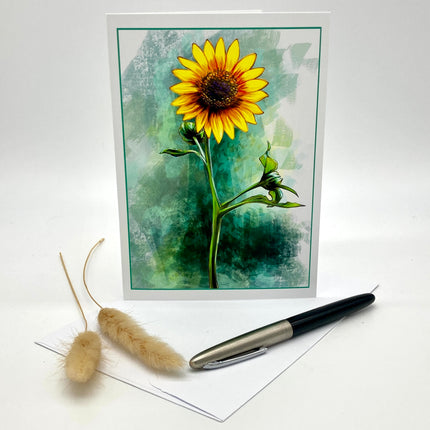 A greeting card featuring a watercolor-style yellow sunflower on a textured green and teal background, displayed upright with an envelope and a black-and-silver pen on a white surface. Two dried bunny tail grass stems are placed to the left.