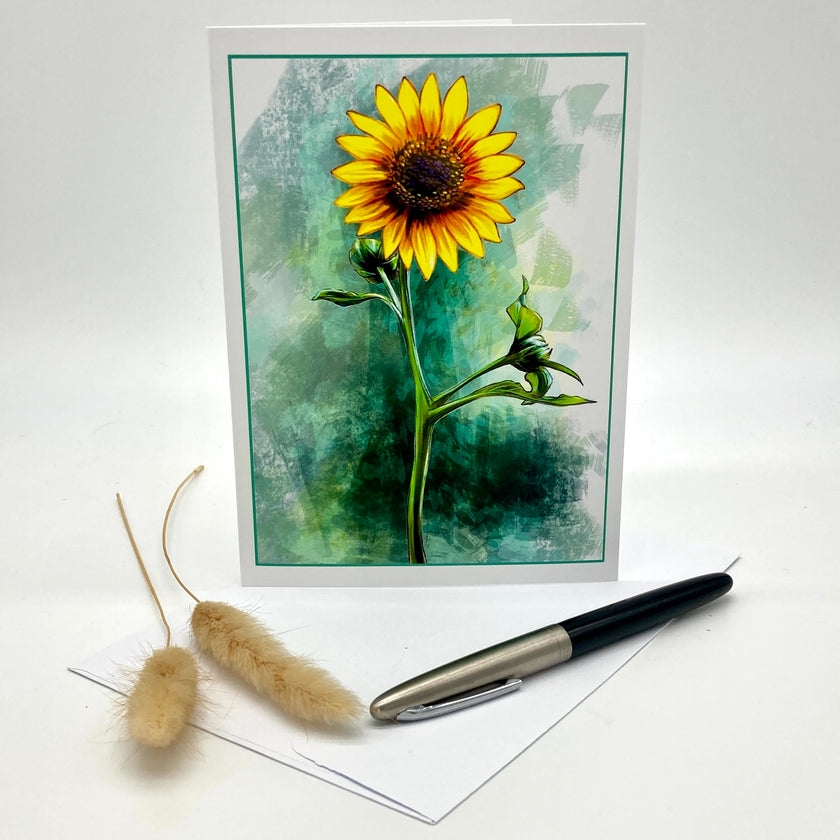 A greeting card featuring a watercolor-style yellow sunflower on a textured green and teal background, displayed upright with an envelope and a black-and-silver pen on a white surface. Two dried bunny tail grass stems are placed to the left.