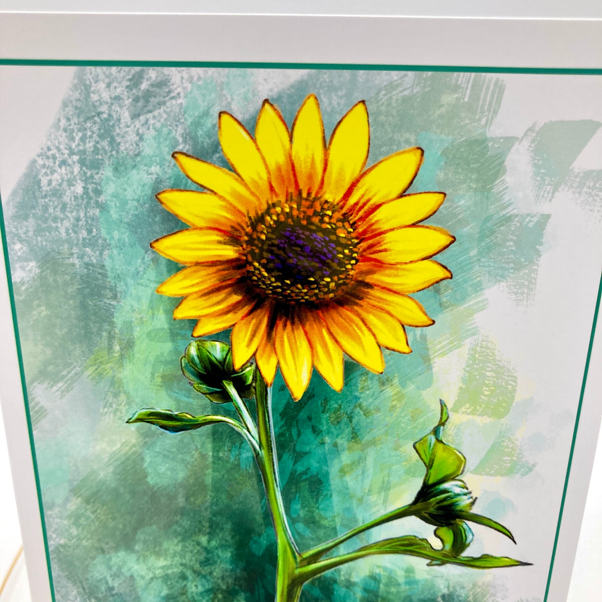 A close-up of a square art print featuring a bright yellow sunflower with green leaves and stems against a textured teal and white background.
