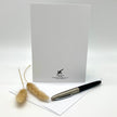 A white stationery card or envelope standing upright on a white surface, with a black and silver pen resting on the front and two dried beige grass stems beside it. The card has a small black logo and text near the bottom.