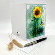 A white greeting card standing upright on a white surface, featuring a printed watercolor-style sunflower illustration on the front. A silver and black pen and two dried grass stems rest beside the card.