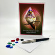 A birthday greeting card featuring a whimsical illustration of a bird wearing a party hat, displayed upright on a white surface with a pen, an envelope, and small colorful gem-like accessories in front.