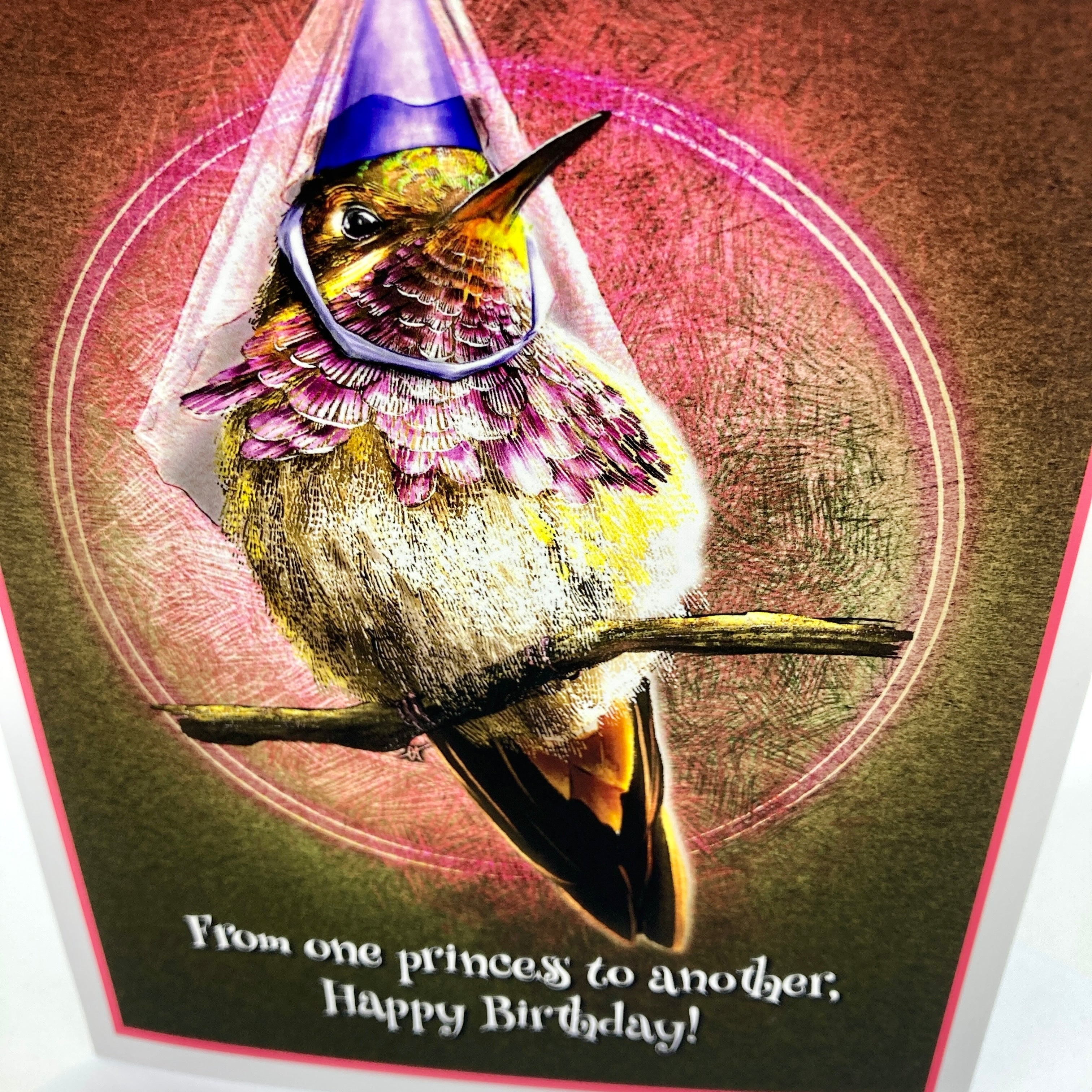A close-up of a birthday card featuring an illustrated hummingbird wearing a purple party hat and a silver ring, perched on a branch against a textured brown and pink background. The card reads, 'From one princess to another, Happy Birthday!' in white script at the bottom.