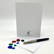 A white greeting card set with a black and silver pen and small colorful gemstone charms, photographed on a clean white background.