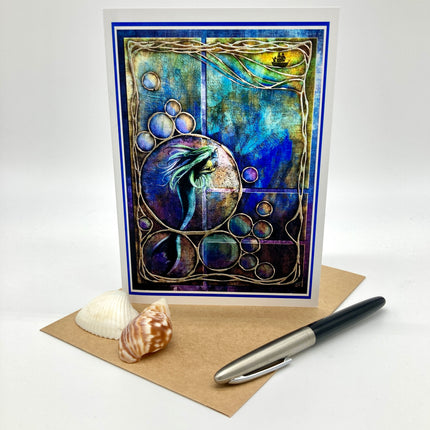 A square art print or greeting card with a mermaid illustration in blue, purple, and gold tones, displayed upright on a kraft paper envelope with two seashells and a black-and-silver pen on a white studio background.