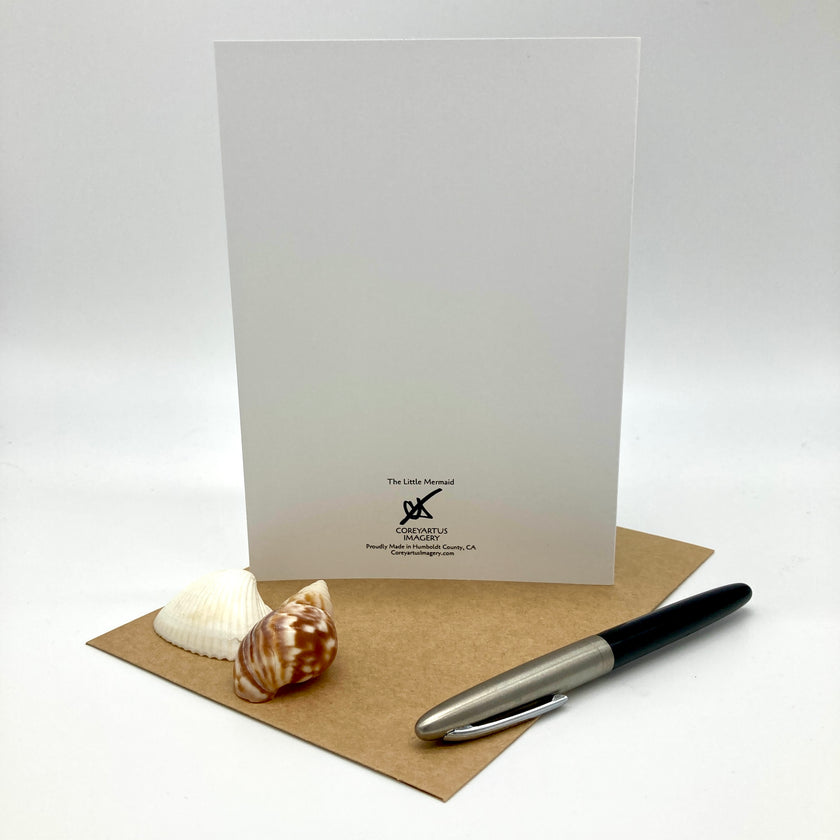 A white greeting card with a black logo and text stands upright on a brown kraft paper envelope, styled with a white seashell and a black-and-silver pen on a light gray background.