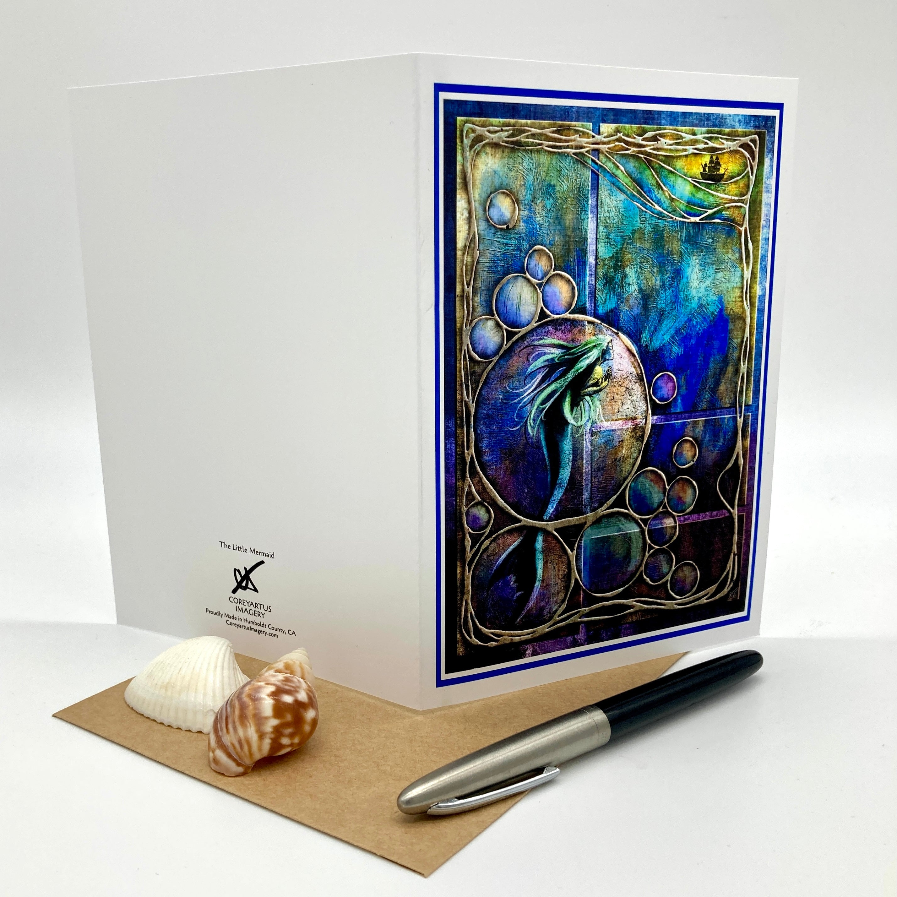 A white greeting card with a colorful mermaid artwork on the front, displayed with a brown envelope, two seashells, and a black-and-silver pen on a white studio background.