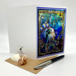 A white greeting card with a colorful mermaid artwork on the front, displayed with a brown envelope, two seashells, and a black-and-silver pen on a white studio background.