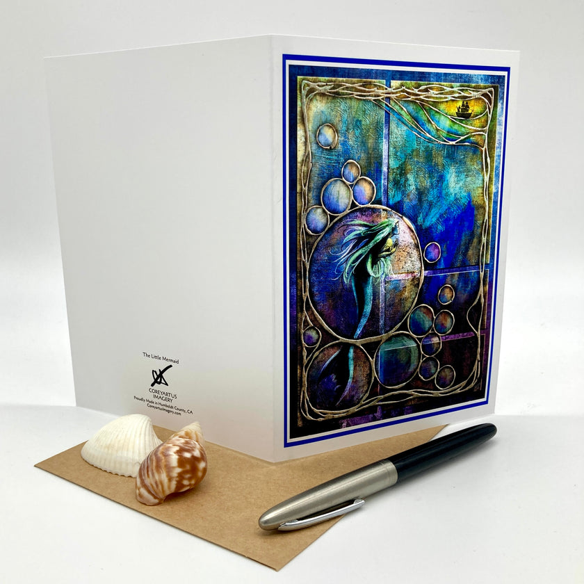 A white greeting card with a colorful mermaid artwork on the front, displayed with a brown envelope, two seashells, and a black-and-silver pen on a white studio background.