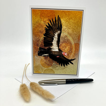 A greeting card featuring a black bird with white wing details and a pink head against a textured gold and brown background, displayed upright on a white surface with a pen and two dried grass stems in front.