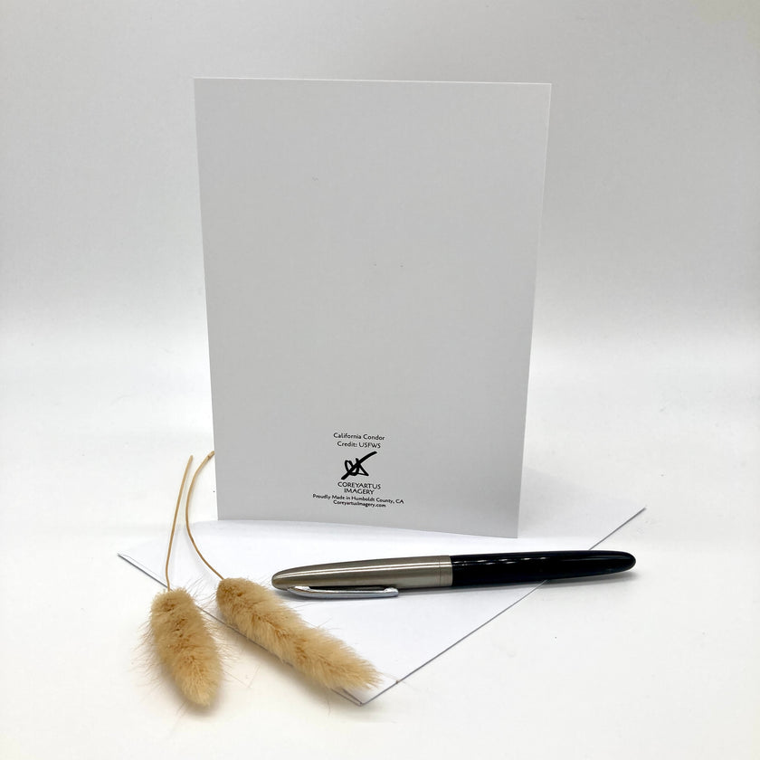 A white greeting card or notepad standing upright on a white surface, with a black and silver pen resting across the lower corner and two dried beige grass stems in front. Small printed branding is visible near the bottom center of the card.