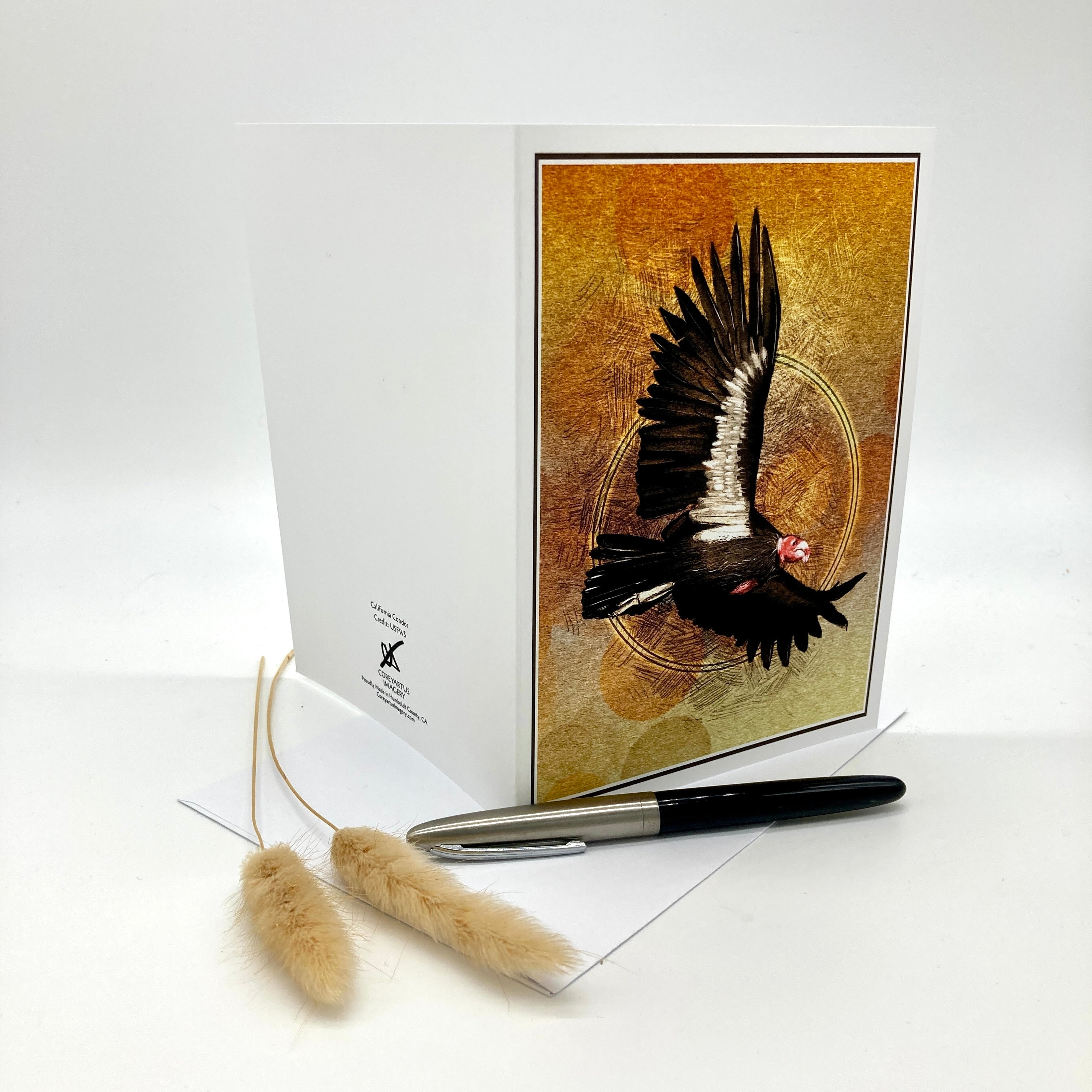 A white greeting card standing upright, featuring a printed illustration of a black bird with white wings over a textured gold background. A black and silver pen and two dried beige grass stems are placed in front on a white surface against a light gray studio background.