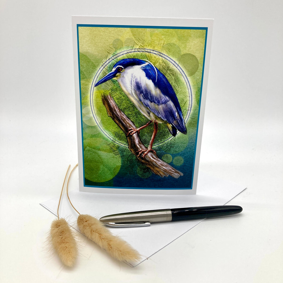 A square greeting card featuring a detailed illustration of a blue and white bird perched on a branch against a green and blue abstract background. The card is displayed upright on a white surface with a black pen and two dried bunny tail stems in front.