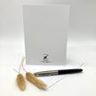 A white greeting card with a black logo and text at the bottom, displayed upright on a white surface with a silver and black pen in front and two dried beige grass stems beside it.
