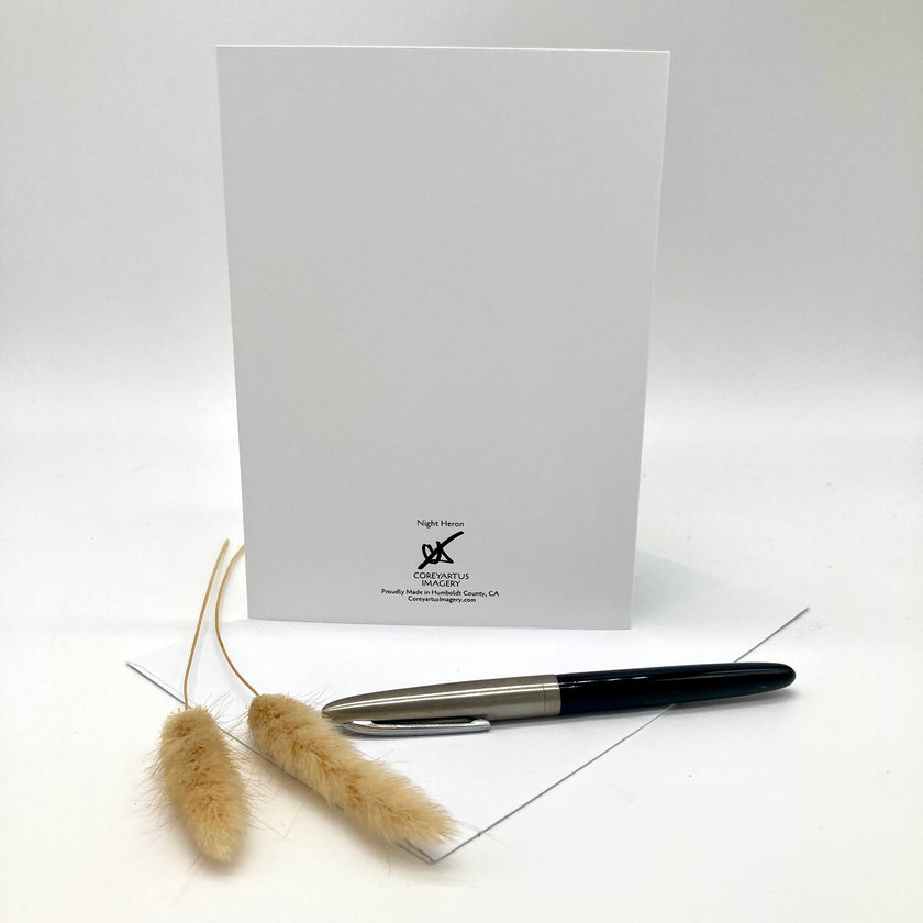 A white greeting card with a black logo and text at the bottom, displayed upright on a white surface with a silver and black pen in front and two dried beige grass stems beside it.