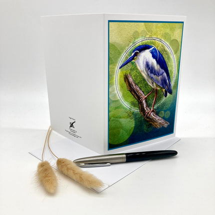 A white greeting card with a printed illustration of a blue and white heron perched on a branch against a green and blue background. A matching white envelope and a black-and-silver pen are placed in front on a white surface, with two dried grass stems to the left.