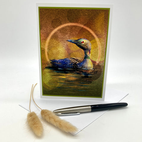 A vertical greeting card featuring a painted loon in water with a glowing circular halo, displayed upright on a white surface with a pen and two dried grass stems in front.