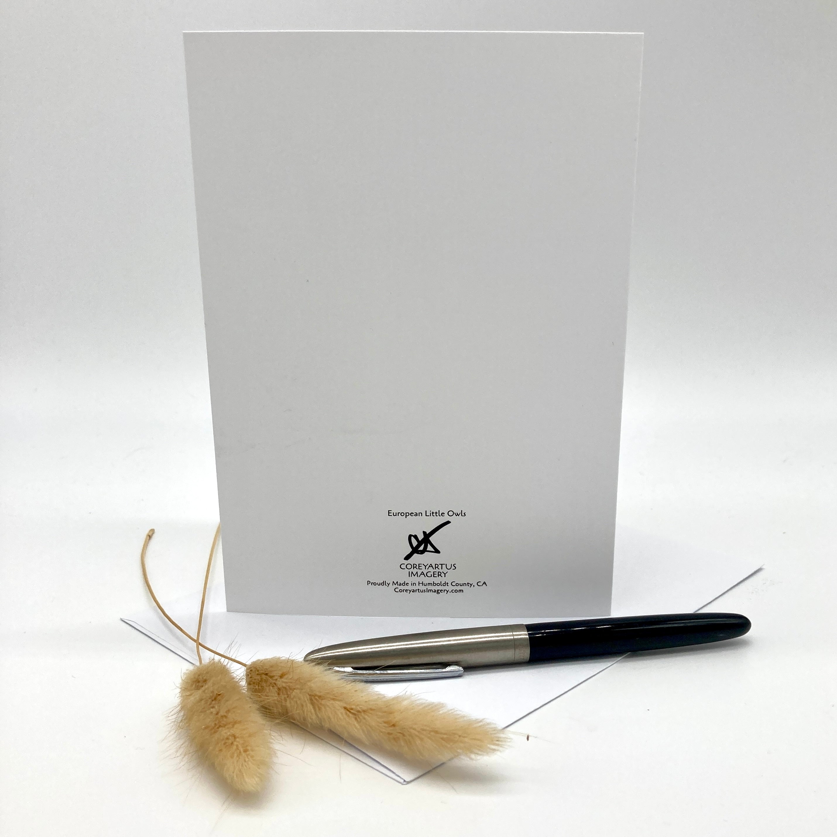 A white greeting card with black printed branding at the bottom, displayed upright on a white background with a black and silver pen and two dried beige grass stems in front.