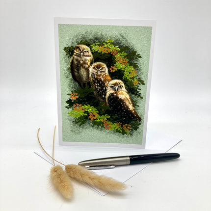 Product photo of a greeting card posed with complimentary props on a white background. Card features three burrowing owls with different expressions staring straight at the viewer against aa green paper background.