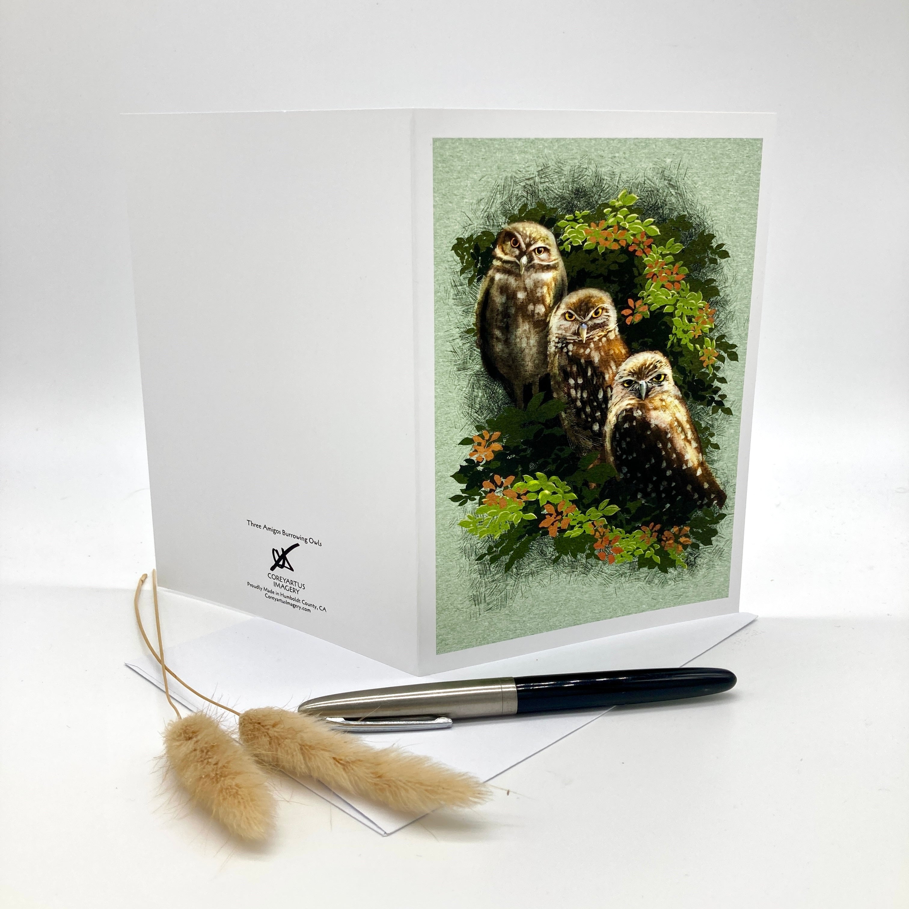 A white greeting card standing upright on a white surface, featuring an illustration of three owls among green leaves and small orange flowers on the front. The back of the card is visible on the left, with a small printed logo and text.