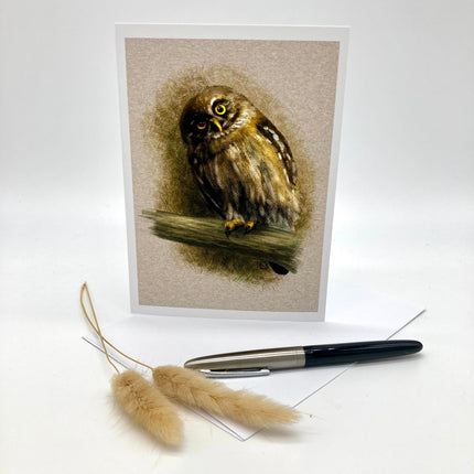 A vertical art print or greeting card featuring a painted owl perched on a branch, shown standing upright on a white surface with a pen and dried grass stems in front.