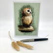 A vertical greeting card featuring a detailed illustration of a brown owl perched on a branch against a textured sage green background. The card is displayed upright on a white surface, with a black and silver pen and two dried pampas grass stems placed in front.