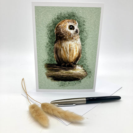 A vertical greeting card featuring a detailed illustration of a brown owl perched on a branch against a textured sage green background. The card is displayed upright on a white surface, with a black and silver pen and two dried pampas grass stems placed in front.