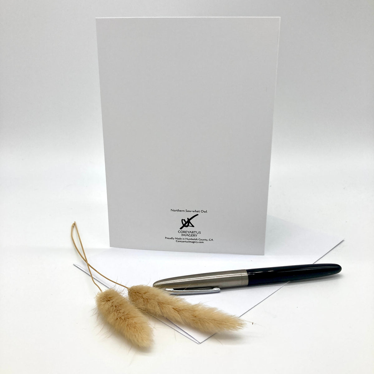 A white greeting card with the 'Harper's Bazaar Club' logo printed at the bottom, displayed with a black and silver pen and dried pampas grass on a white surface against a light gray background.
