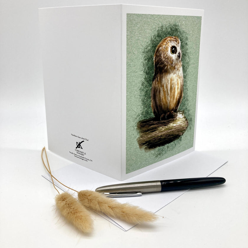 A white greeting card featuring a detailed illustration of a brown owl perched on a branch against a muted sage green background. The card is displayed on a white surface next to a black and silver pen and two dried wheat stalks.