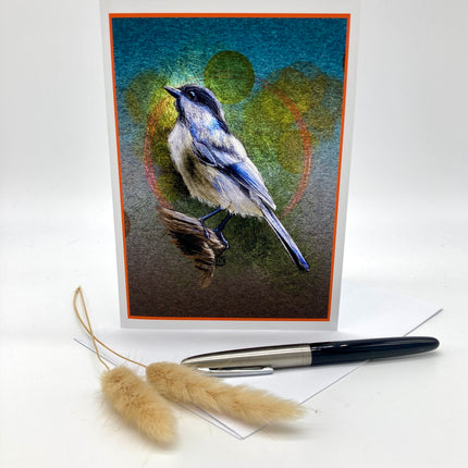 A square greeting card featuring a painted bird perched on a branch, displayed upright on a white surface with a pen and dried grass stems in front. The card has an orange border and a textured blue, green, and brown background.