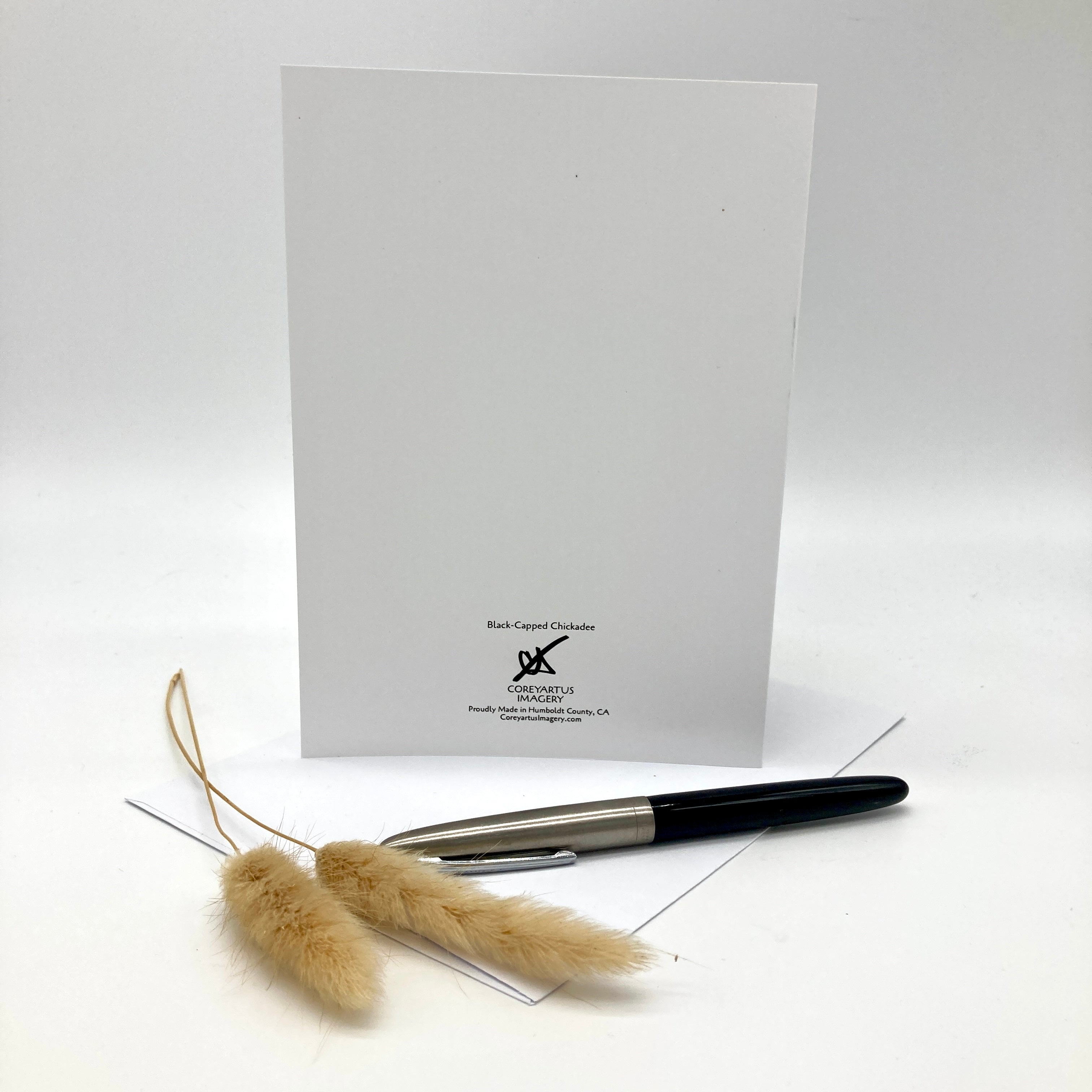 Backside of a greeting card, posed with a spring of wild grasses and a black ink pen on a white envelope against a white background.