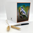 A folded greeting card displayed upright on a white surface, featuring a colorful illustration of a bluebird on the front. A white envelope and a black-and-silver pen sit in front, with dried bunny tail grass stems placed near the card.