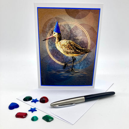 Photograph of a Greeting Card featuring a Marbled Godwit in a blue conical party hat. Beside the card are plastic rhinestones and an ink pen. 