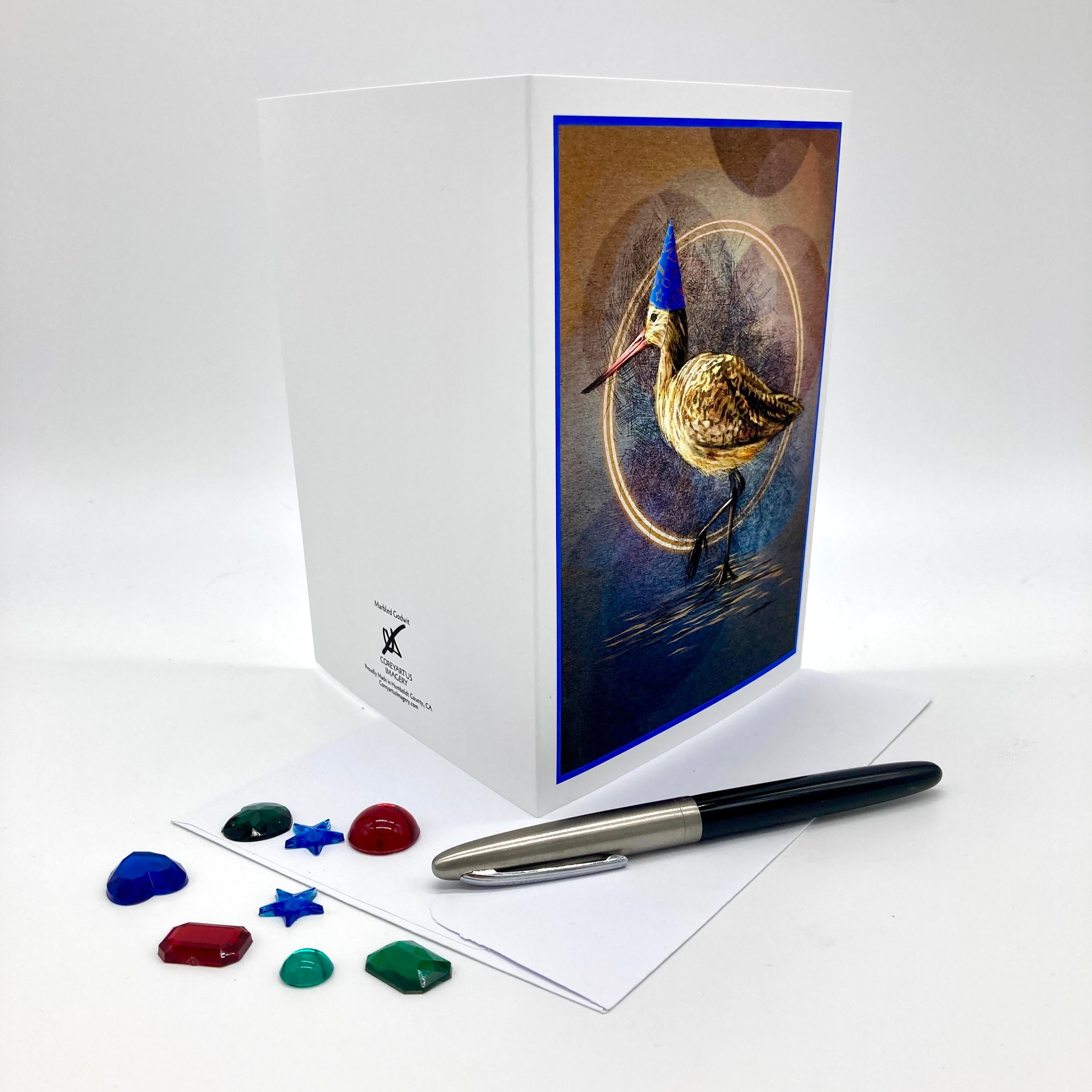 Photo of the backsides of a Marbled Godwit birthday card splayed out open, posed with plastic rhinestones and an ink pen.