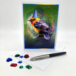 Photo of a greeting card posed with plastic rhinestones and an ink pen. The card features a flame-colored tanager in a blue conical party hat against a green and purple background.